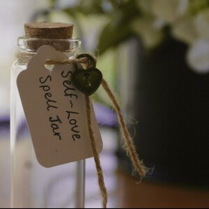 Pu&ograve; includere: Un piccolo barattolo di vetro con un tappo di sughero e un'etichetta bianca che dice "Self-Love Spell Jar". Un piccolo lucchetto a forma di cuore &egrave; attaccato all'etichetta con uno spago.