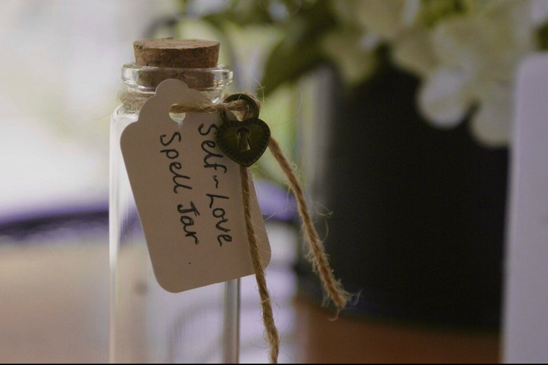 Pu&ograve; includere: Un piccolo barattolo di vetro con un tappo di sughero e un'etichetta bianca che dice "Self-Love Spell Jar". Un piccolo lucchetto a forma di cuore &egrave; attaccato all'etichetta con uno spago.