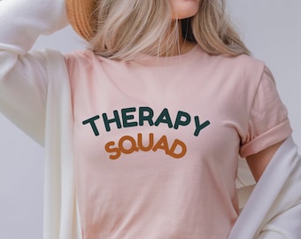 May include: A light pink t-shirt with the words "THERAPY SQUAD" in green and orange lettering. The shirt is being worn with a white cardigan and a straw hat. The shirt is made of a soft material.