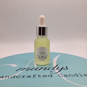 May include: A clear glass bottle with a silver cap and a white dropper. The bottle is filled with a light yellow liquid. The label on the bottle says "Jasmine". The bottle is sitting on a light blue surface with the text "mandys handcrafted candle" printed on it.