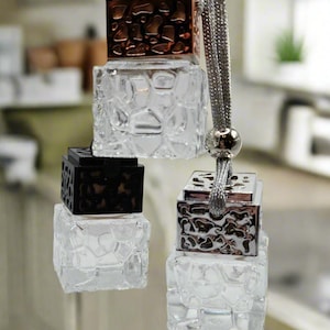 May include: Three car air fresheners with different colored tops. The top air freshener has a copper colored top, the middle air freshener has a black top, and the bottom air freshener has a silver top. All three air fresheners have a clear glass bottle with a textured surface.