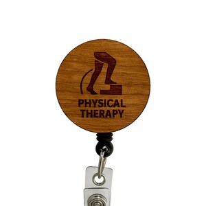 Puede incluir: Un portatarjetas de madera con un diseño circular. El diseño presenta las piernas de una persona subiendo una escalera, con las palabras "PHYSICAL THERAPY" debajo. El carrete tiene un cordón negro, un cierre de metal y un clip de plástico transparente.