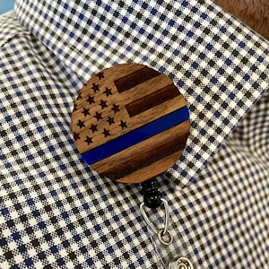 Puede incluir: Un porta credencial de madera con un diseño de bandera estadounidense y una línea azul. El porta credencial está unido a un carrete retráctil para credenciales.
