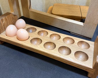May include: A wooden egg holder with a capacity for twelve eggs. The holder is made of light-colored wood and has rounded depressions to hold the eggs. Three light-colored eggs are in the holder. The holder is part of a larger wooden structure.