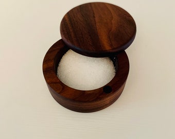 May include: A round wooden salt cellar with a lid. The cellar is made of dark wood and has a small hole in the side. The lid is also made of dark wood and has a smooth surface. The cellar is filled with white salt.