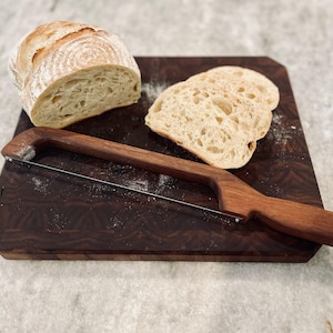 May include: A wooden bread board with a bread knife and two slices of sourdough bread. The board has a dark brown wood grain pattern.