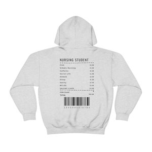 Könnte beinhalten: Ein grauer Kapuzenpullover mit einem schwarz-weißen Beleg auf der Rückseite. Der Beleg lautet "Nursing Student" und listet Artikel wie "Figs", "Simple Nursing", "Caffeine", "Social Life", "HOKAS", "Sleep", "Sanity", "NCLEX" und "Saving Lives" auf. Der Gesamtbetrag beträgt 32,95 €.