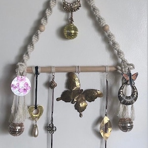 Puede incluir: Un tapiz de macramé de pared con una barra de madera y varios artículos decorativos colgados, incluyendo una mariposa, un corazón, una bola de discoteca y varios pendientes.