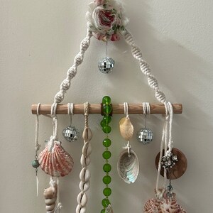 Puede incluir: Un tapiz de macramé blanco de pared con un acento de tela floral, una barra de madera y varios elementos colgantes, incluyendo conchas marinas, cuentas y bolas de discoteca.