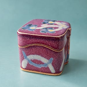 May include: A square ceramic lidded box with a speckled magenta glaze. The lid features blue and yellow floral motifs, while the base displays a light blue curved pattern. The container has a textured finish and a distinct, slightly wavy rim.