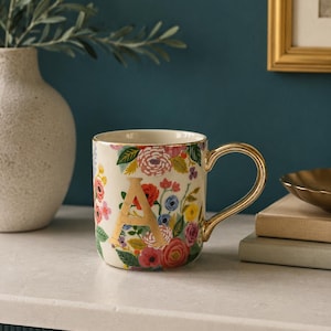 May include: A cream-colored ceramic mug featuring a vibrant floral pattern of pink, red, yellow, and blue flowers. The mug displays a large, gold-colored letter A on the front, a gold-rimmed top edge, and a shiny gold-toned handle.