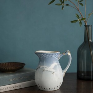 May include: A porcelain creamer pitcher featuring a light blue gradient top with a scale pattern and a white base. The body is decorated with a painted white bird in flight, accented with gold trim on the handle and rim, standing on a dark surface.