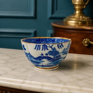 May include: A ceramic bowl featuring a blue and white transferware pattern depicting a landscape with a bridge, trees, and figures. The bowl has a scalloped rim and a base, both accented with a thin gold-colored trim along the edges.