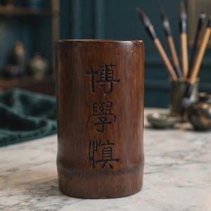 May include: A cylindrical dark brown wooden brush holder with a natural bamboo texture. The front surface features three vertically aligned, deeply carved Chinese characters that read 博學慎. The container has a smooth, polished finish.