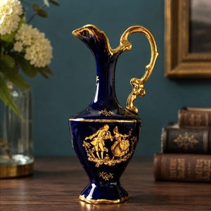 May include: A deep blue porcelain jug featuring an ornate gold handle and a gold-painted scene of two figures in historical attire dancing. The rim, base, and decorative accents are finished in gold, contrasting against the dark blue glaze.