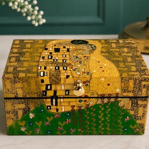 May include: A rectangular wooden box featuring a reproduction of Gustav Klimt's The Kiss painting. The exterior displays gold, black, and green patterns with a central couple embracing. A small, round, metallic gold knob is centered on the front lid.