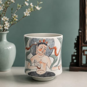 May include: A cylindrical ceramic cup featuring a hand-painted illustration of a seated figure with a halo, rendered in shades of orange, grey, and black against a white background. The figure is depicted with flowing ribbons and traditional attire.