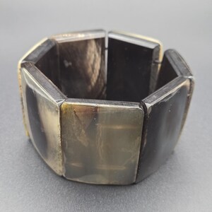 May include: A chunky bracelet with a geometric design. The bracelet is made of dark, rectangular segments with a polished finish. The color palette includes shades of black, brown, and tan, creating a natural, earthy aesthetic. The bracelet is designed to be worn around the wrist.