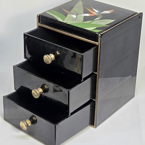 May include: A black lacquer jewelry chest with three tiered drawers and gold-toned knobs. The top surface features a painted illustration of a bird of paradise flower with green leaves against a black background, framed by gold-colored edges.