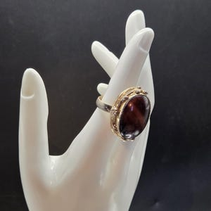 May include: A silver and gold ring with a large, oval, reddish-brown stone. The ring has a decorative gold setting around the stone and small accent stones. The ring is displayed on a white hand against a black background.