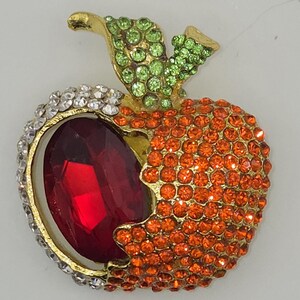 May include: A jeweled apple brooch with a gold-tone stem and leaves. The apple is encrusted with orange rhinestones, a large red gemstone, and a border of white rhinestones. The leaves are green.