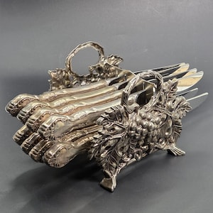 May include: A set of silver-toned metal knives with ornate, embossed handles featuring floral and leaf motifs, held within a decorative silver-toned metal stand. The stand is detailed with a prominent grape cluster and vine leaf design on both sides.