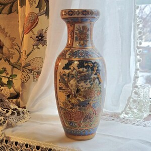 May include: A tall, ornate ceramic vase with a flared neck and base. The vase features a detailed, colorful design with birds, flowers, and a landscape scene. Gold accents and trim enhance the intricate patterns. The vase is approximately 12 inches tall.