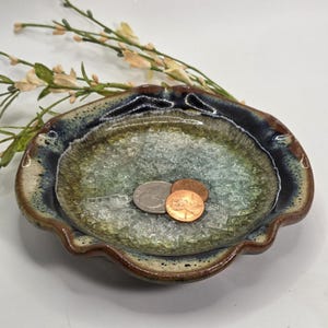 May include: A handmade ceramic dish with a scalloped edge, featuring a blue, green, and brown glaze. The dish contains clear, crushed glass and three coins. Decorative sprigs of flowers are in the background.