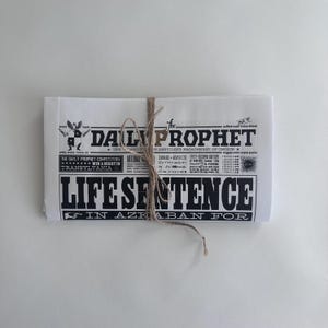 May include: A folded white newspaper replica titled The Daily Prophet with black text and graphics. The headline reads Life Sentence In Azkaban For. The paper is tied with a thin brown twine string across the centre, featuring various small printed details.