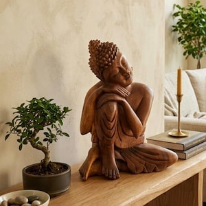 Könnte beinhalten: Eine geschnitzte Holzskulptur einer sitzenden Buddha-Figur mit einer strukturierten, braunen Oberfläche. Die Figur ist in einer ruhenden Pose dargestellt, mit einem über das Knie gelegten Arm und geneigtem Kopf, was die Details hervorhebt.