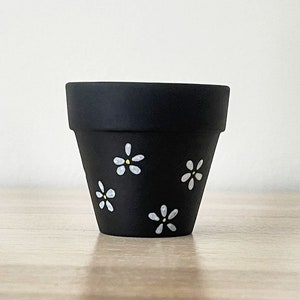 May include: A small, black ceramic plant pot decorated with white daisy motifs. The pot has a gently sloping form and is adorned with hand-painted white daisies with yellow centres. Suitable for small plants or succulents.