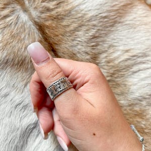 May include: A silver thumb ring with a repeating pattern of arrows and a stylized cow head design.