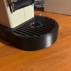 May include: Close-up of an espresso machine with a cream-colored body and a black drip tray. The machine has chrome accents and a black handle. The drip tray has a circular indentation and a slatted design.
