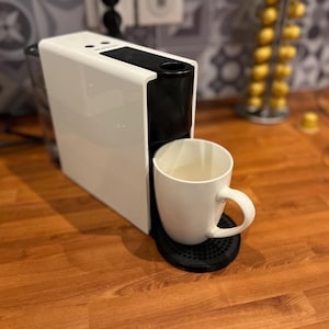May include: A white and black espresso machine with a white mug on a black base. The machine is on a wooden surface. The mug is filled with a light-coloured liquid.