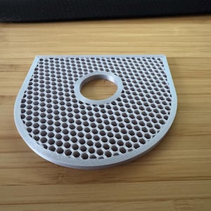 Puede incluir: Un filtro de color plateado en forma de U, con un patrón de panal de abeja y un agujero redondo en el centro.