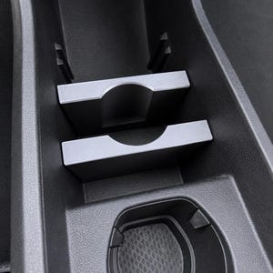 May include: A black car console with a built-in cup holder and a tray for small items. The console has a textured surface and a matte finish. The cup holder has a textured bottom.