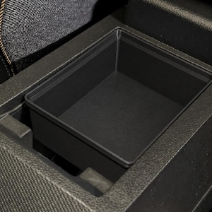 Op de afbeelding: Een rechthoekig opbergbakje van zwart kunststof, ontworpen voor de middenconsole van een voertuig. Het bakje heeft een gladde, platte bodem en verticale wanden en past precies in het uitgespaarde compartiment van de zwarte, getextureerde console.