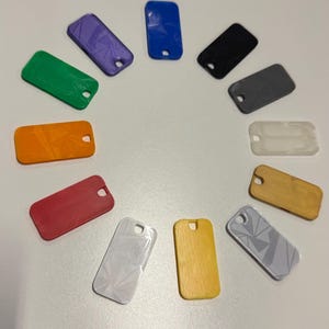 Può includere: Una collezione di 12 piccoli portachiavi rettangolari in vari colori e motivi, tra cui bianco, grigio, nero, blu, verde, arancione, rosso e giallo. I portachiavi sono disposti in cerchio su una superficie bianca.