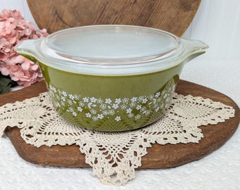 May include: A round olive green glass casserole dish with a clear glass lid and integrated side handles. The exterior features a repeating white floral pattern consisting of small blossoms and leaves encircling the middle section.