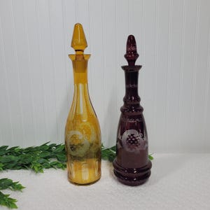 May include: Two vintage glass decanters with etched designs. The decanter on the left is amber glass with a white etched design. The decanter on the right is a deep burgundy color with a white etched design.