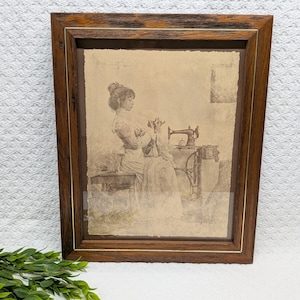 May include: A framed sepia-toned print depicts a woman sewing at a treadle sewing machine. The artwork is encased in a dark brown wooden frame with a gold inner trim. The print shows the woman in a long dress, seated, with her hands near the machine.