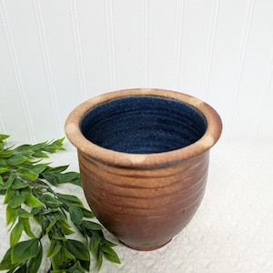 May include: A handmade, brown ceramic pot with a dark blue interior. The pot has a textured, ridged exterior and a light brown rim. The design is simple and rustic, suitable for holding plants or as a decorative item.