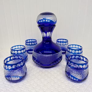May include: A cobalt blue glass decanter with a matching stopper and six glasses. The set features intricate cut glass designs with white accents. The decanter and glasses are arranged on a white surface, showcasing the glassware's elegant details.