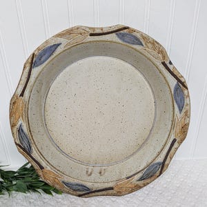 May include: A speckled, beige ceramic pie plate with a decorative leaf design around the rim. The leaves are in shades of brown, tan, and blue. The plate has a slightly scalloped edge.