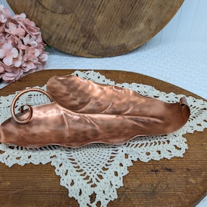 May include: A hammered copper tray shaped like an elongated leaf with a curled stem handle on one end. The metallic surface features a textured, dimpled finish that reflects light, creating a warm, reddish-brown appearance throughout the piece.