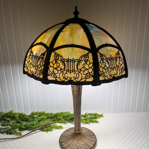 May include: An antique table lamp with a stained glass shade. The shade features a colorful, mosaic-like design with yellow, orange, and blue hues, framed by black metal filigree. The lamp has a bronze-colored base and stem.