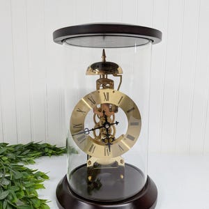 May include: A vintage brass and glass clock with Roman numerals. The clock mechanism is visible through the open face. The clock is encased in a clear glass dome with a dark brown wooden base and top.