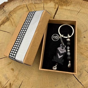 May include: A keychain featuring a black and gray wolf charm, a circular black charm with the white letter F, and a strand of black and silver beads attached to a silver metal ring, presented inside a brown cardboard gift box with a checkered ribbon.