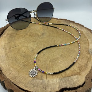 Op de afbeelding: Een zwarte kralenbrilkoord met een zilveren zonnebloembedel. De ketting is gemaakt van kleine zwarte kralen met kleurrijke accenten. De ketting ligt op een houten oppervlak.