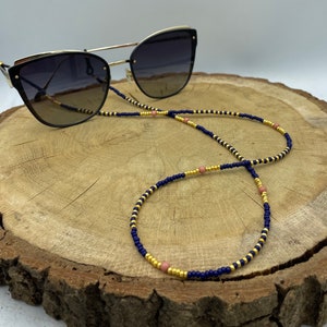 Op de afbeelding: Een navyblauwe en gouden kralenketting voor brillen met roze kralen. De ketting ligt op een houten oppervlak.